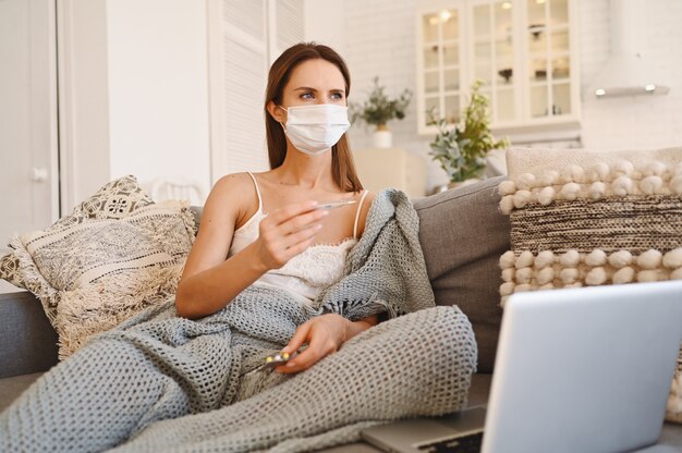 Donna malata che indossa la maschera di protezione del viso, seduto sul divano con il computer portatile e tenendo termometro e pillole durante l'autoisolamento di quarantena domestica.