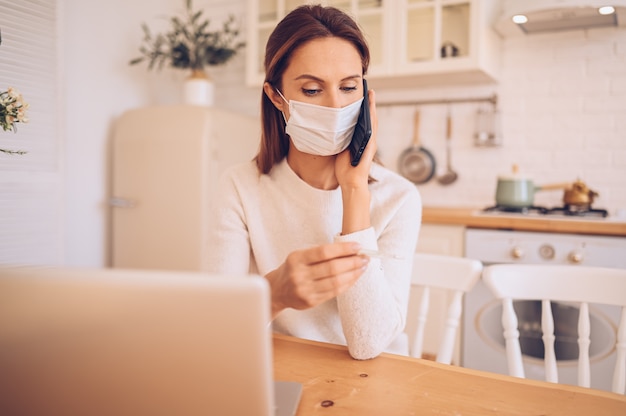 donna malata che indossa una maschera medica, parlando di smartphone e lavorando su un laptop in cucina a casa durante l'isolamento di quarantena pandemia di Covid-19