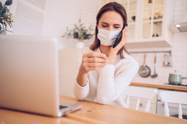 donna malata che indossa una maschera medica, parlando di smartphone e lavorando su un laptop in cucina a casa durante l'isolamento di quarantena pandemia di Covid-19