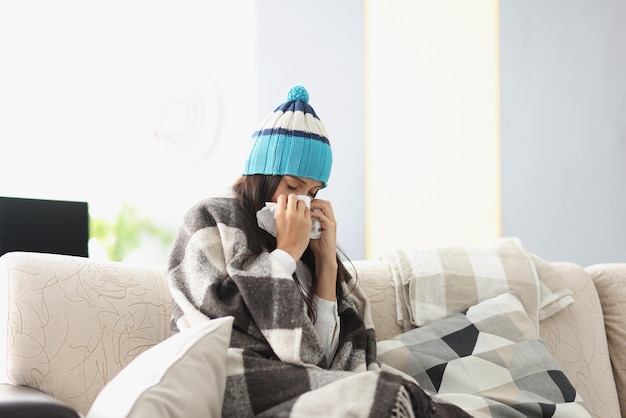 Donna malata con un cappello caldo che si soffia il naso nel tovagliolo di carta a casa