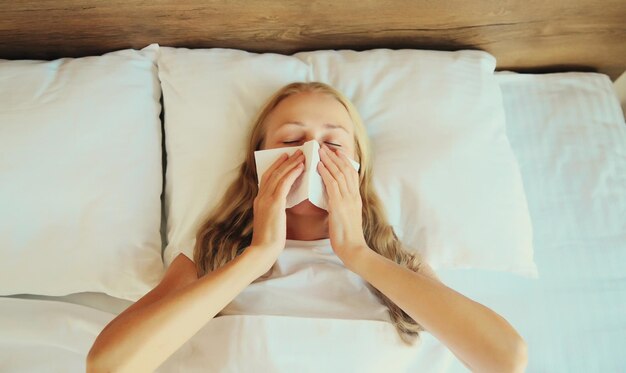 Donna malata sconvolta che starnutisce soffiando il naso usando un fazzoletto sdraiata a letto a casa