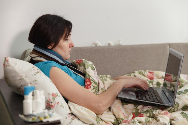 Donna malata sdraiata a letto a casa che lavora al computer portatile.