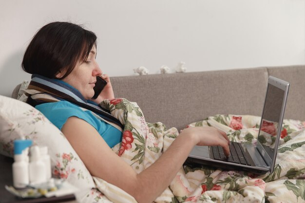 Donna malata sdraiata a letto a casa, parlando al telefono e lavorando al computer portatile.