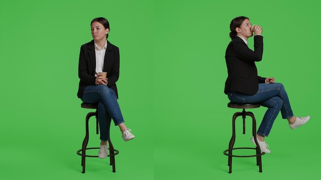 Donna manager che beve una tazza di caffè prima di lavorare in ufficio, seduto su una sedia in tuta. Impiegato aziendale che si gode una bevanda alla caffeina in studio, in posa su uno schermo verde per tutto il corpo.