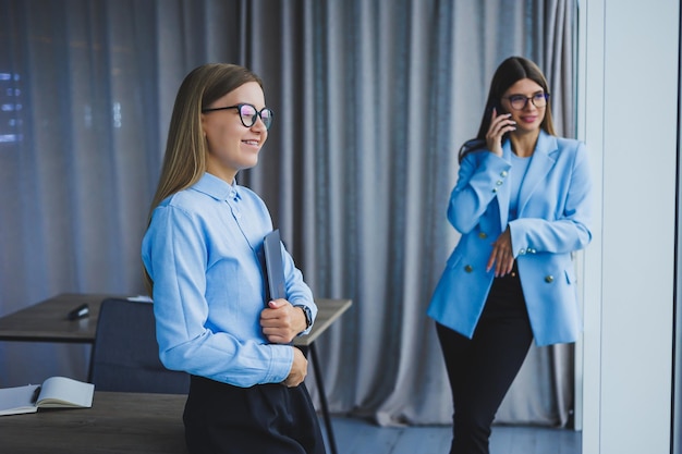 Donna manager con occhiali classici sorridente durante l'orario di lavoro in ufficio donna europea positiva in camicia blu scrivania con laptop lavoro a distanza Un collega è sullo sfondo il fuoco selettivo