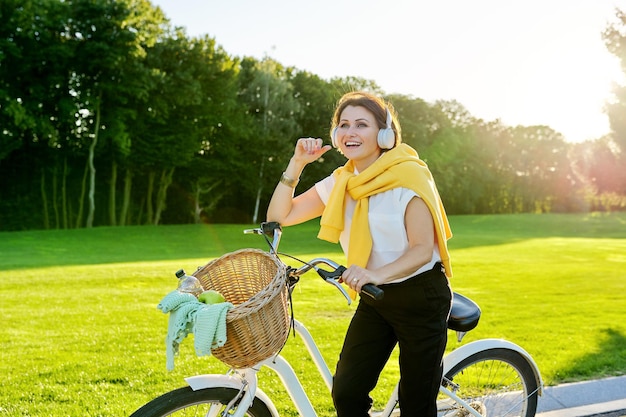 Donna matura all'aperto con la bici. Bella donna di mezza età felice in cuffia con la bicicletta nel parco. Riposo attivo, stile di vita attivoRiposo attivo, età, gioia e felicità delle persone anziane
