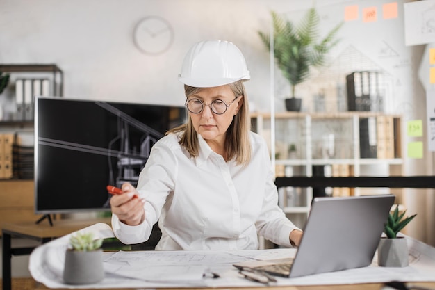 Donna matura architetto o costruttore femminile seduto sulla scrivania sul posto di lavoro in un ufficio moderno