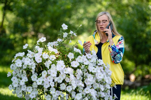 Donna matura bionda vestita in modo brillante che parla sullo smartphone nel concetto di comunicazione online del giardino