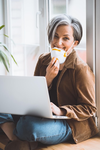 Donna matura che abbassa la maschera fp e sorride guardando la telecamera mentre lavora il computer portatile seduto su win...