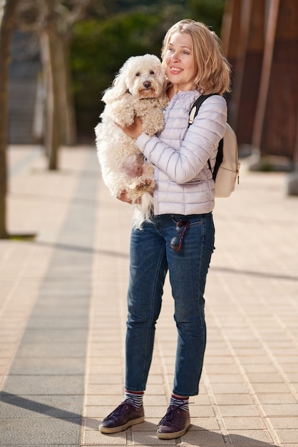 Donna matura che cammina con soffice cane bianco nella città estiva.