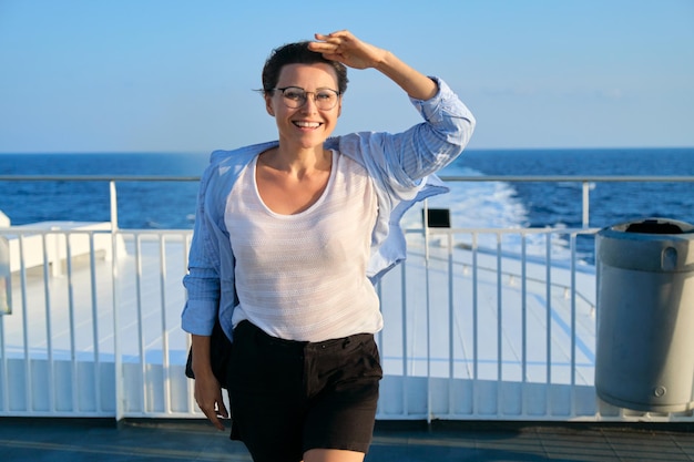 Donna matura che gode della crociera in mare. Femmina in camicia blu sul ponte della nave guardando il paesaggio marino, sfondo del tramonto del mare del cielo. Concetto di vacanza, viaggio, trasporto marittimo, turismo e trasporto