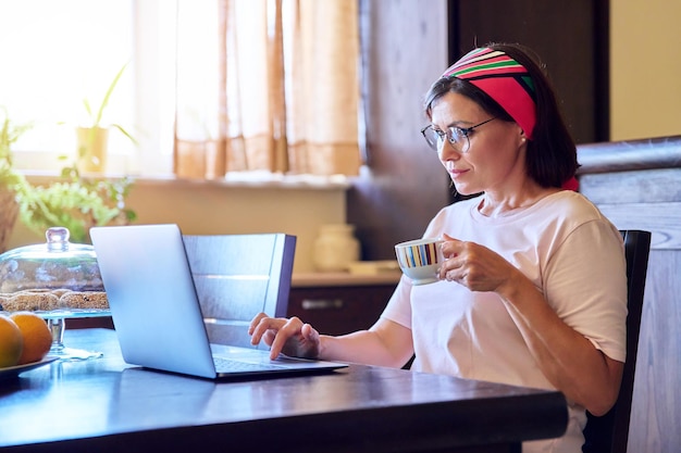 Donna matura che si rilassa a casa al tavolo del soggiorno con un computer portatile e una tazza di caffè