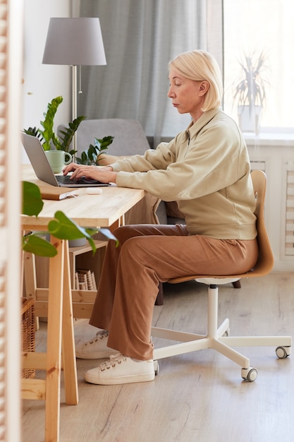 Donna matura che si siede al suo posto di lavoro a casa e che lavora al computer portatile