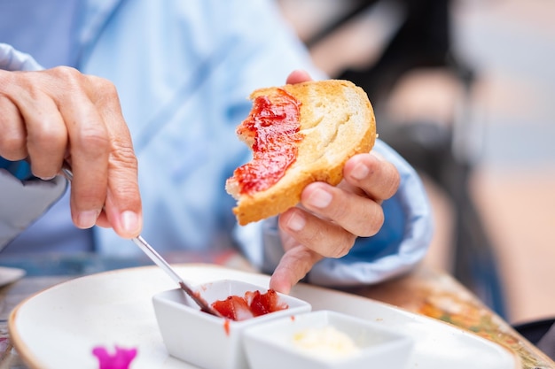 Donna matura che tiene una fetta di pane tostato mettendo sopra la marmellata di fragole mentre si siede a un tavolo da caffè all'aperto per la colazione