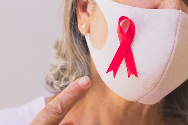 Donna matura con maschera pandemica e nastro rosa isolato su bianco. Immagine del mese di sensibilizzazione sul cancro al seno.