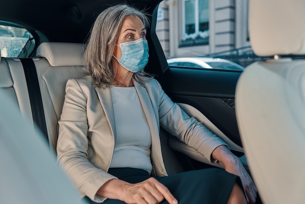 Donna matura con maschera protettiva che guarda attraverso una finestra mentre è seduta sul sedile posteriore dell'auto.