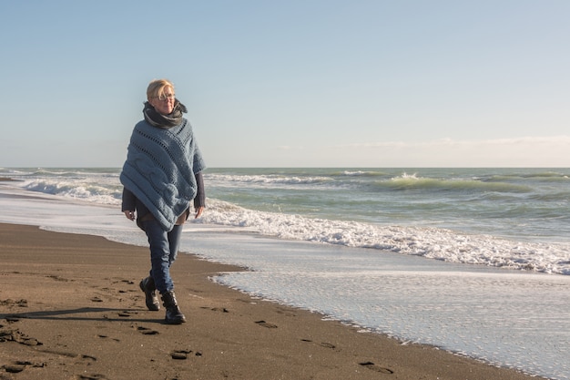 Donna matura con poncho che cammina in riva al mare in autunno