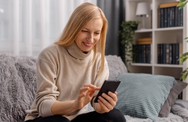 Donna matura felice con i capelli biondi che tocca lo schermo dello smartphone moderno mentre si siede su un divano grigio accogliente Concetto di gadget e stili di vita delle persone