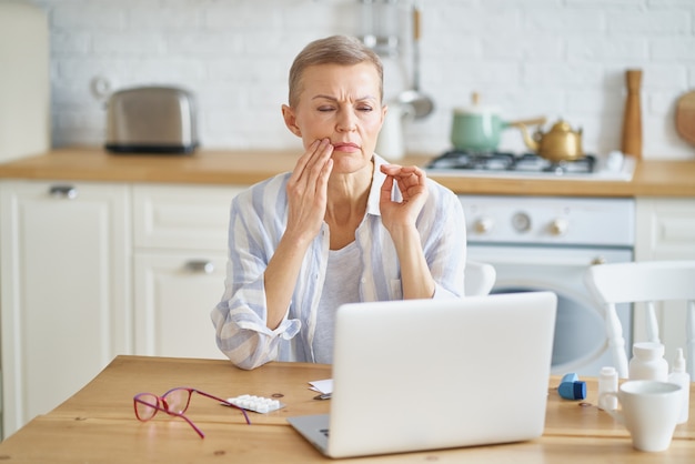 Donna matura frustrata che soffre di mal di denti mentre lavora online da casa