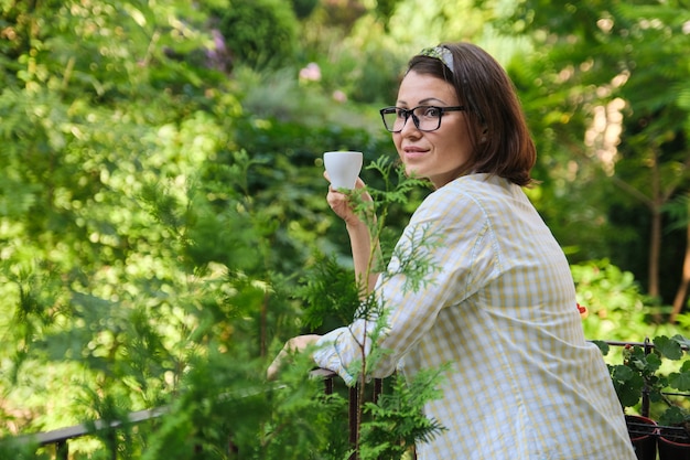 Donna matura in piedi sul balcone aperto, femmina femmina con una tazza di caffè da solo