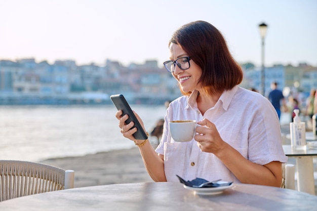 Donna matura in un caffè all'aperto sul marciapiede sul lungomare con una tazza di caffè utilizzando lo smartphone