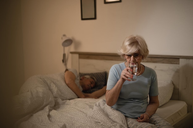 Donna matura pensierosa che beve un bicchiere d'acqua mentre è seduta sul letto di notte Suo marito sta dormendo sullo sfondo