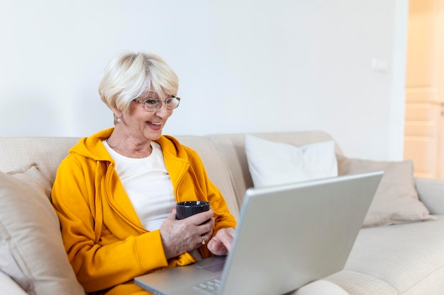 Donna matura più anziana che utilizza app per laptop wireless navigando in Internet sedersi sul divano Donna sorridente di mezza età che fa una videochiamata con i suoi amici o familiari Donna anziana con una tazza di caffè o tè