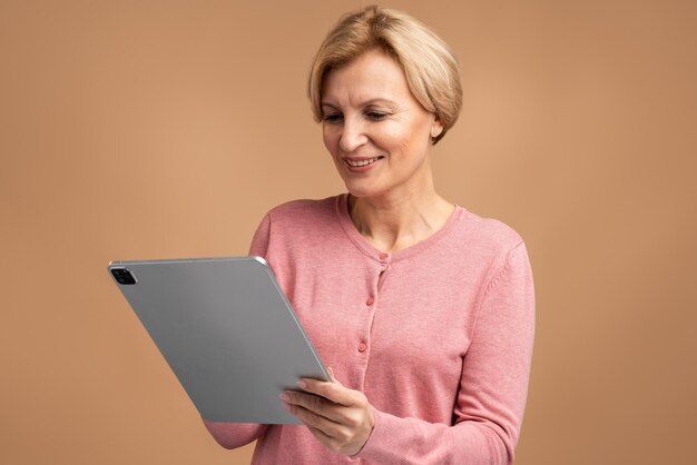 Donna matura positiva che lavora online, utilizza tablet per controllare i social network, guardando sorridendo lo schermo. Colpo dello studio dell'interno isolato su fondo beige