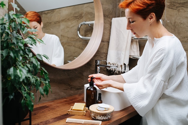 Donna matura premurosa che pompa il sapone ecologico sulle mani per lavarle sopra un lavandino. È in piedi davanti allo specchio in un bagno.