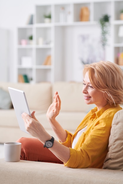 Donna matura riposante felice in abbigliamento casual rilassante sul divano mentre beve il tè e comunica tramite chat video in tablet a casa