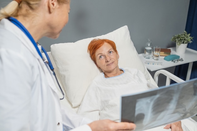 Donna matura sdraiata sul letto e ascoltando il medico che parla dell'immagine a raggi X nel reparto