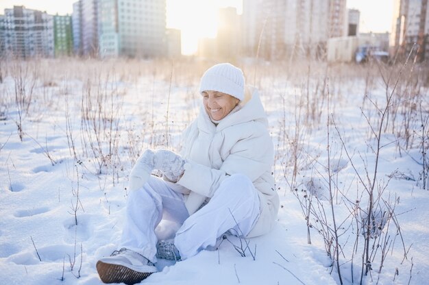 Donna matura senior anziana felice in outwear caldo bianco che gioca con la neve nell'inverno soleggiato all'aperto