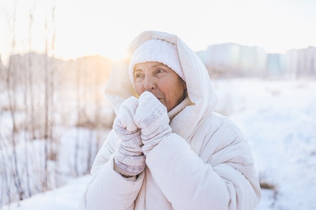 Donna matura senior anziana felice in outwear caldo bianco che gioca con la neve nell'inverno soleggiato all'aperto