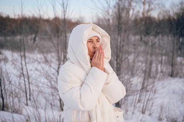 Donna matura senior anziana felice in outwear caldo bianco che gioca con la neve nell'inverno soleggiato all'aperto