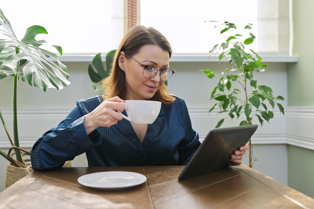 Donna matura sorridente che fa colazione a casa in pigiama con una tazza di tè seduta al tavolo leggendo la tavoletta digitale. Femmina felice dalla posta letta, notizie. Emozioni positive, informazione, vita, 40enni