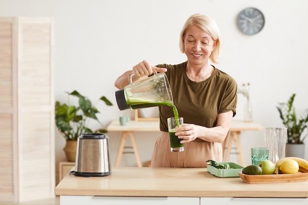 Donna matura sorridente che versa il cocktail della verdura fresca nel bicchiere e lo beve