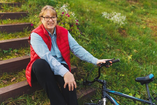 Donna matura sorridente di mezza età che tiene una bicicletta con le mani sull'erba su un campo verde Concetto di vacanza e avventura in campagna estiva o autunnale
