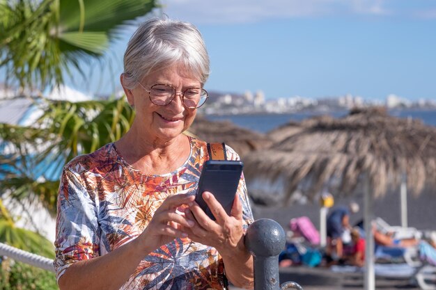 Donna matura sorridente in piedi vicino alla spiaggia con in mano uno smartphone Donna anziana dai capelli bianchi che usa il telefono cellulare godendo di tecnologia e social media