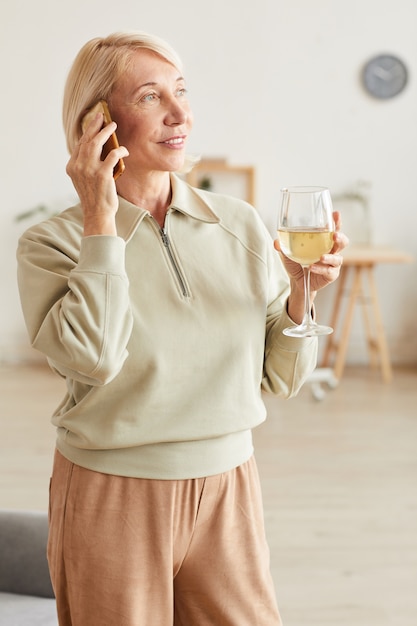 Donna matura tenendo il bicchiere di vino con vino bianco e parlando al telefono cellulare mentre si trovava nella stanza