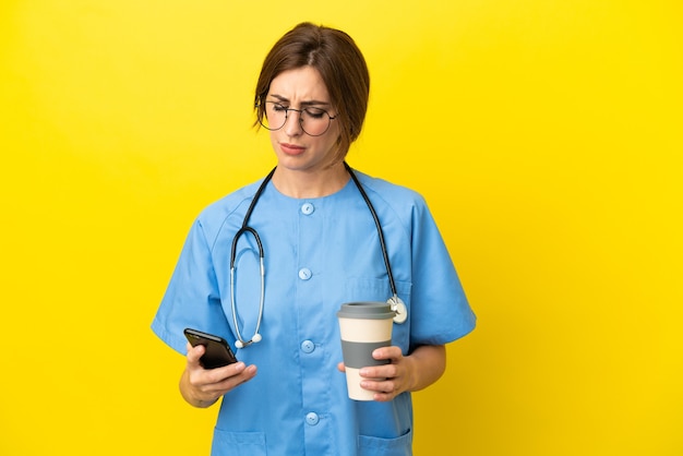 Donna medico chirurgo isolata su sfondo giallo che tiene il caffè da portare via e un cellulare