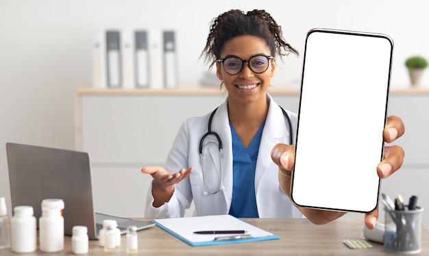 Donna medico nera in uniforme che dimostra un grande smartphone vuoto con schermo bianco