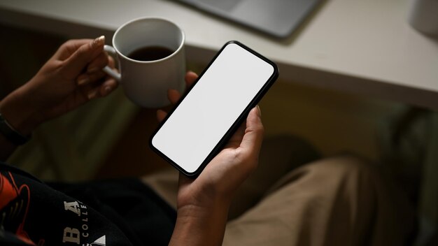 Donna millenaria hipster che tiene una tazza di caffè e uno smartphone mockup con schermo bianco nella moderna stanza buia