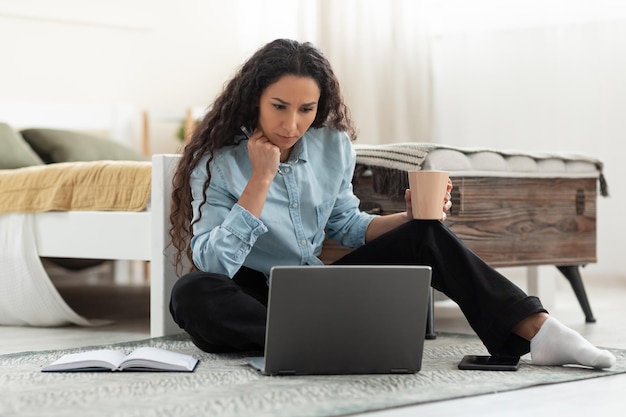 Donna millenaria premurosa che utilizza il pc portatile seduto sul pavimento con il caffè a casa per tutta la lunghezza