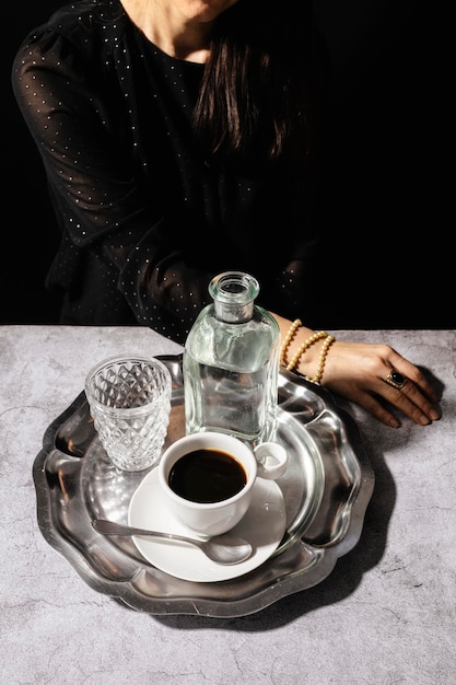 Donna misteriosa che beve caffè nero con bicchiere e brocca d'acqua su un elegante vassoio d'argento con scuro