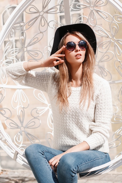 Donna moderna europea giovane hipster in un lussuoso cappello in jeans alla moda in un maglione lavorato a maglia in occhiali da sole in posa seduto su una sedia d'attaccatura vintage bianco metallo in una giornata di sole Ragazza carina.