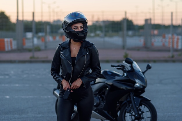 Donna motociclista in giacca di pelle nera e casco integrale si trova vicino alla moto sportiva alla moda. Parcheggio urbano, tramonto in una grande città.