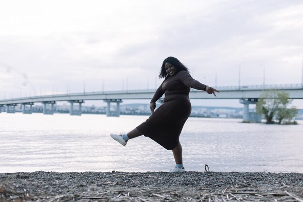 Donna multiculturale dai capelli neri e paffuta afroamericana riccia sorridente e giocosa che scherza vicino alla spiaggia del fiume