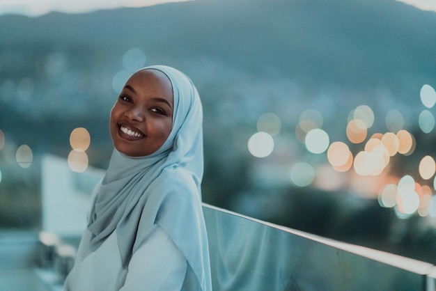 Donna musulmana africana nella notte su un balcone che sorride alla macchina fotografica con le luci del bokeh della città nei precedenti. Foto di alta qualità