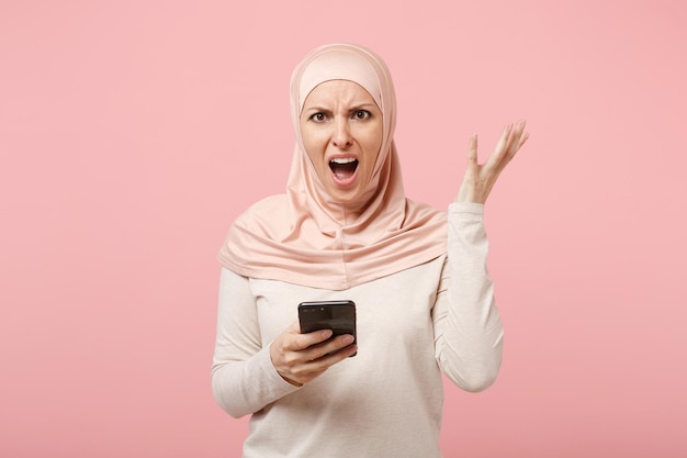 Donna musulmana araba arrabbiata in abiti leggeri hijab in posa isolata su sfondo rosa. Concetto di stile di vita dell'Islam religioso della gente. Mock up spazio di copia. Utilizzando il cellulare digitando un messaggio sms, allargando le mani.