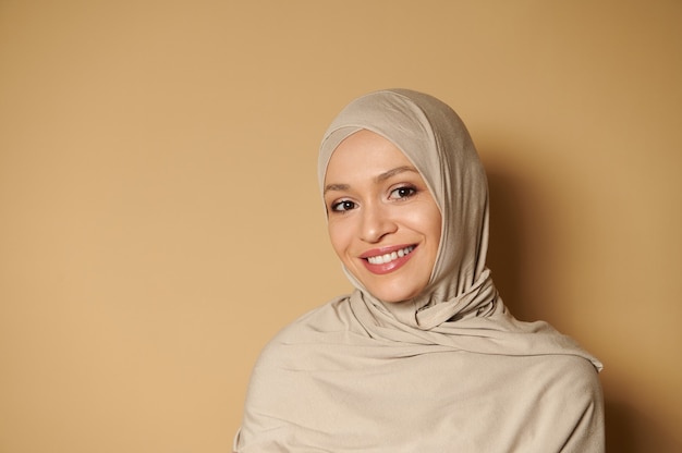 Donna musulmana araba che indossa l'hijab guardando davanti mentre posa con un bel sorriso a trentadue denti su una superficie beige con spazio di copia